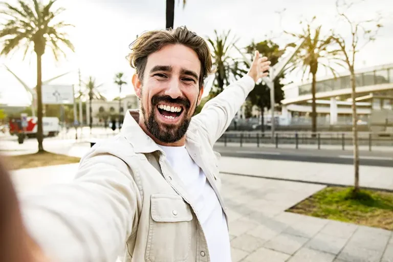 A happy man with a beard and short brown hair is smiling broadly and pointing enthusiastically towards the right, taking a selfie outdoors with palm trees and a city street in the background. The image promotes mental well-being and positive self-expression for Men's Mental Health Awareness Month.