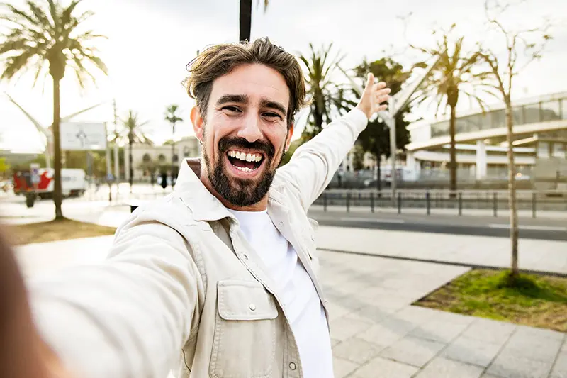 A happy man with a beard and short brown hair is smiling broadly and pointing enthusiastically towards the right, taking a selfie outdoors with palm trees and a city street in the background. The image promotes mental well-being and positive self-expression for Men's Mental Health Awareness Month.