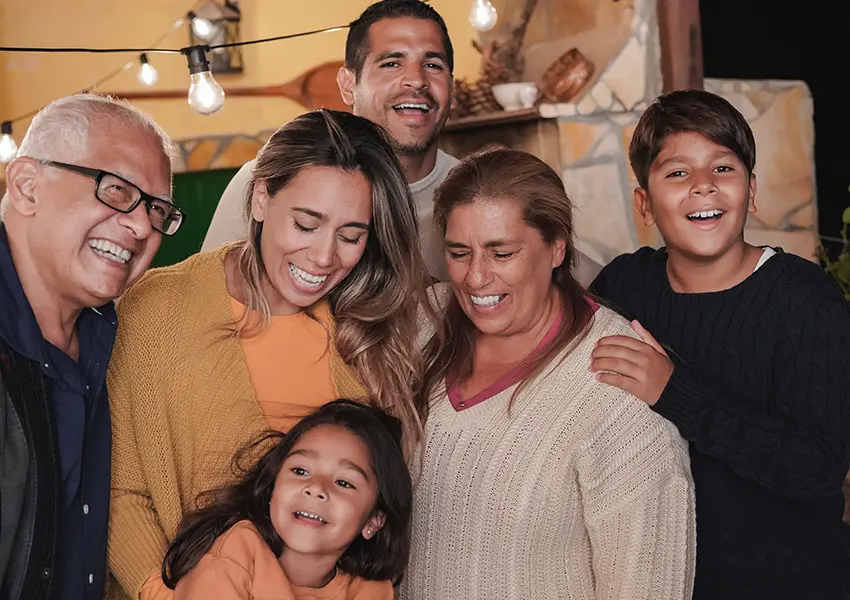 A candid, close-up photo of a multi-generational, Hispanic family of six smiling happily and looking down or toward the center. The image is warm, taken outdoors at night under string lights, conveying joy and connection.