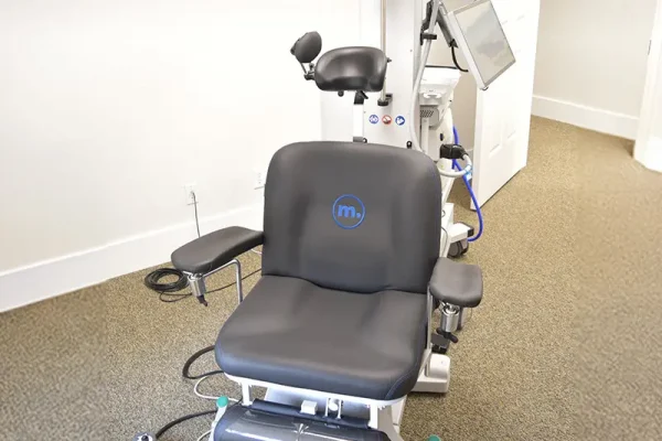A specialized medical chair with a "mo." logo and an attached screen, set up in a bright, white-walled room, representing equipment used for Transcranial Magnetic Stimulation (TMS) therapy.
